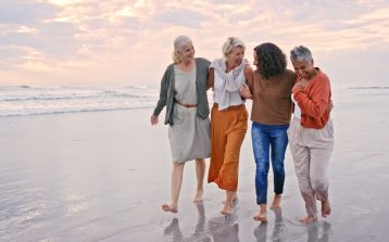 Happy, women and friends on beach, walking and connect on vacation, holiday and bonding together outdoor. Diversity, retirement and senior females talking, stroll and seaside vacation and sunset.