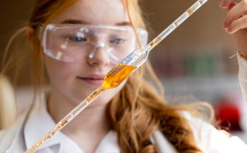 Close-up of a female teenager holding up a pipette filled with an orange substance. She is wearing a lab coat and protective eyewear.